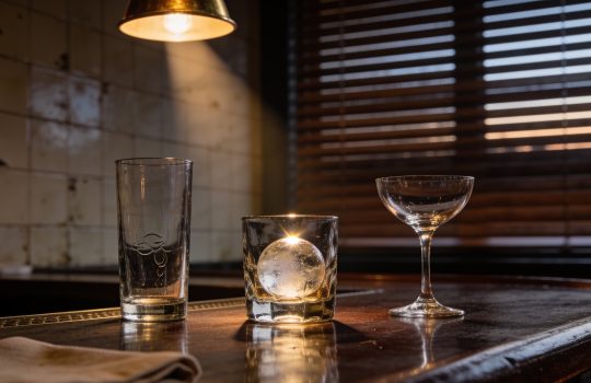 Empty late-evening bar top with three different glasses and an ice sphere in warm lamplight, wooden blind behind